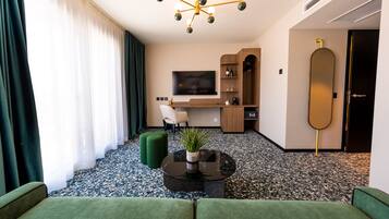 Ein modernes Hotelzimmer mit einem grünen Sofa, einem schwarzen Couchtisch mit Pflanze, einem Fernseher, einem Holztisch und einem großen Spiegel.