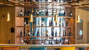 Una zona de bar con estantes de madera que exhiben diversas botellas, un mostrador y asientos con sillas.