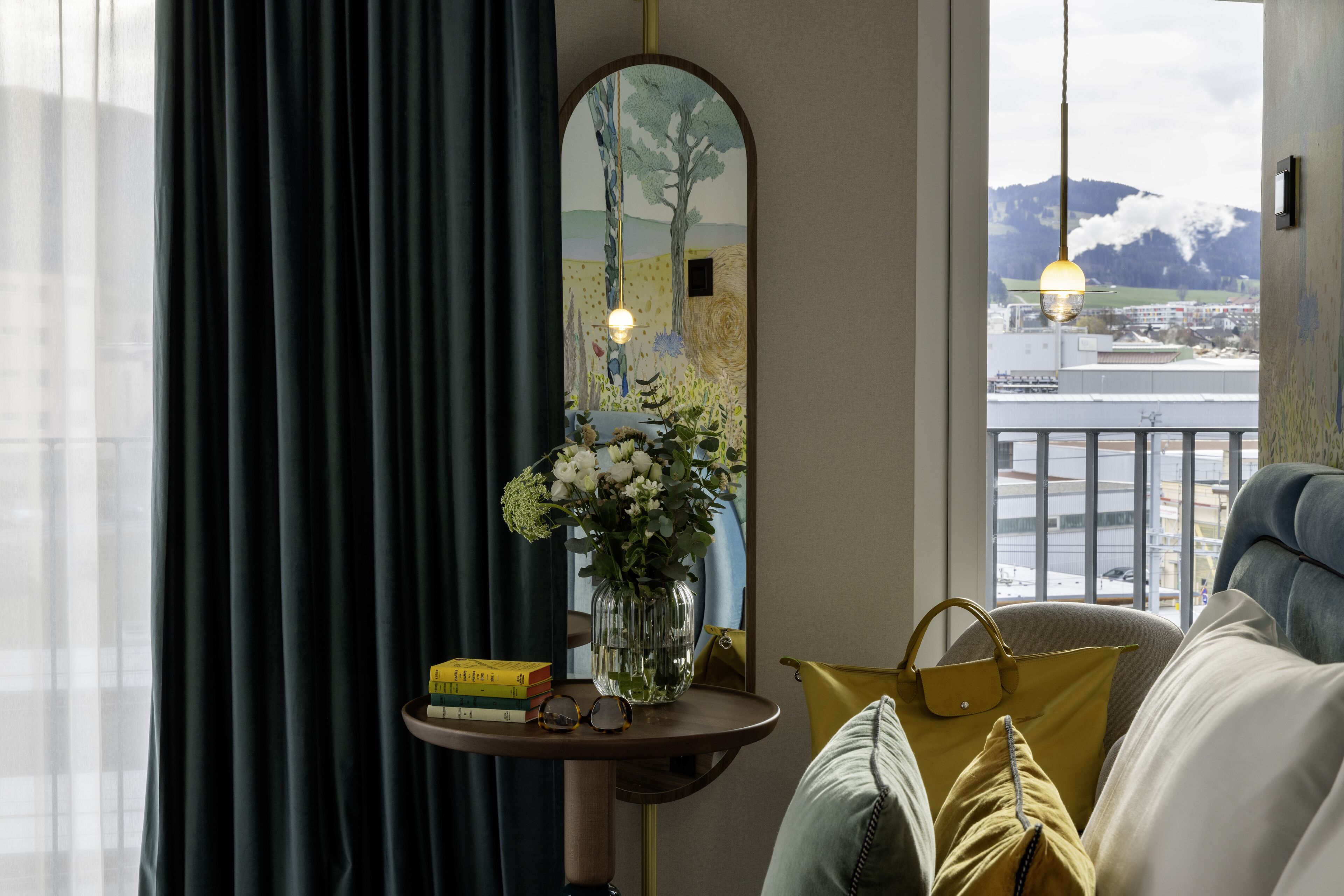 A room with a view of a snowy landscape, a mirror reflecting a tree, a vase with flowers, books, and a handbag on a table.