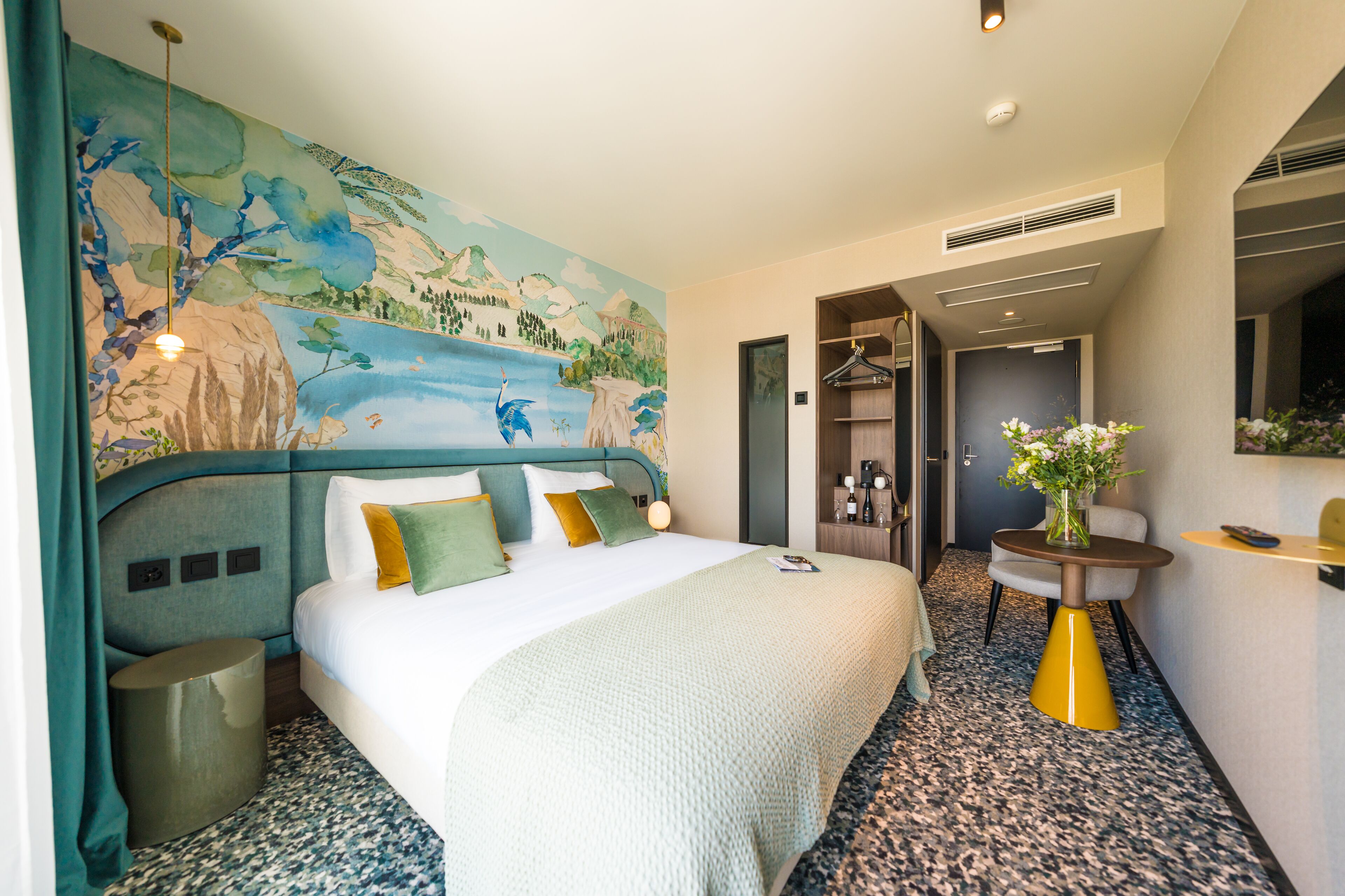 A hotel room with a large bed, a mural on the wall, a small table with a vase, and a mirror.