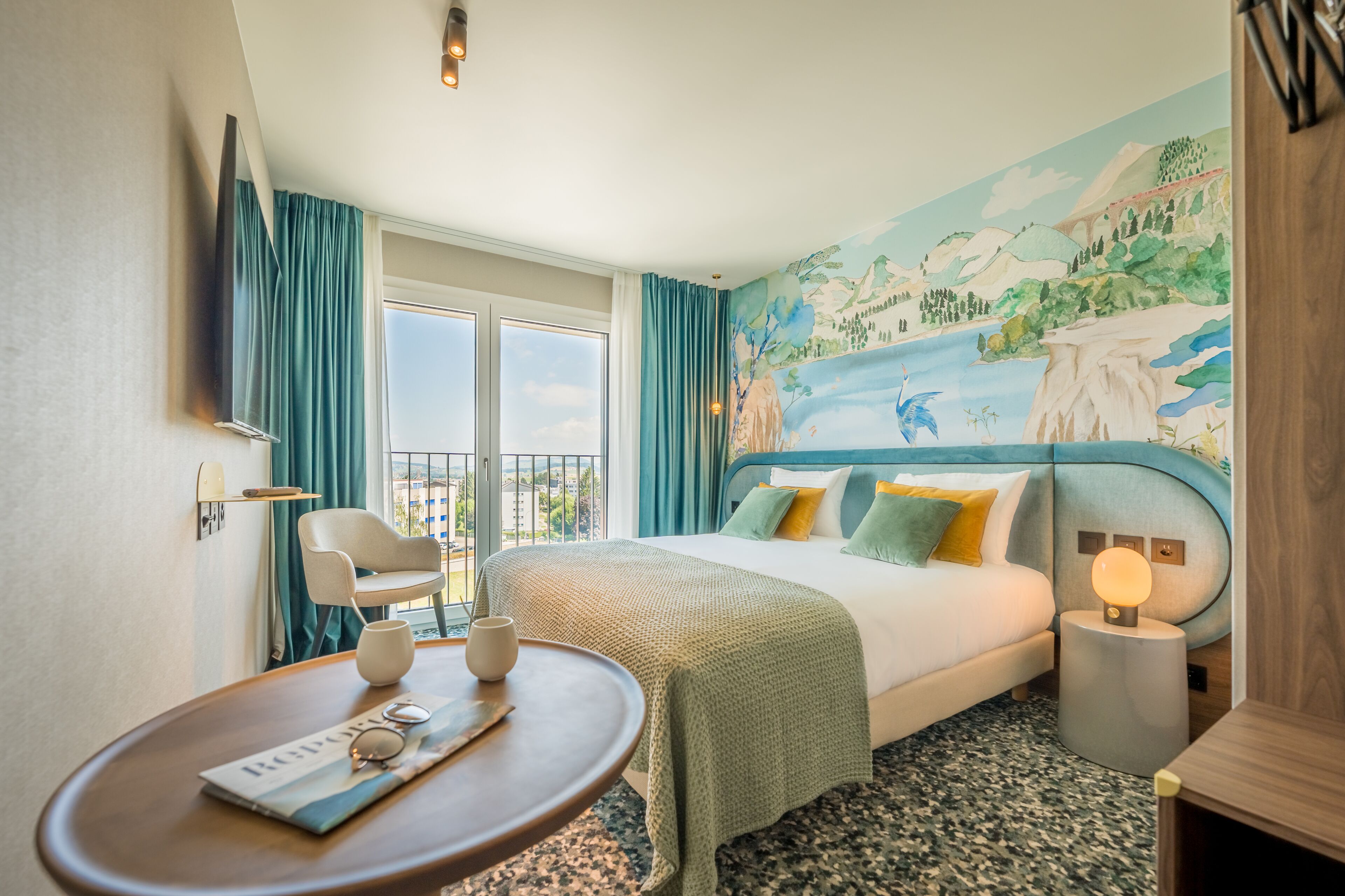 A hotel room with a bed, a round table with a magazine, a chair, a nightstand with a lamp, and a mural on the wall.