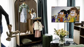 A hotel room with a TV displaying a cartoon, a wooden wardrobe, a hanging chair with a teddy bear, and a green ottoman.