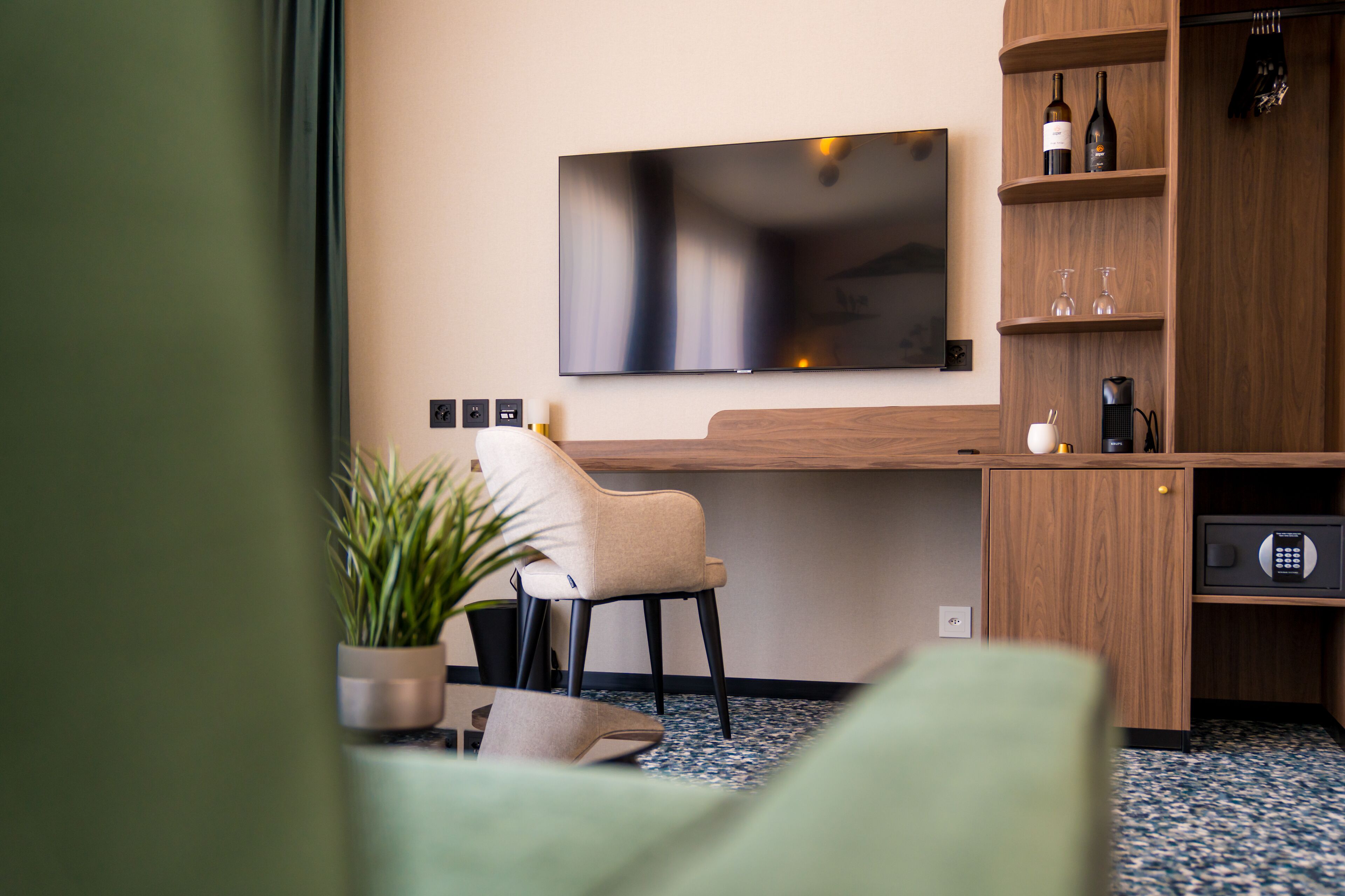 A modern hotel room with a flat-screen TV mounted on the wall, a wooden desk with a chair, a small plant, and a minibar.