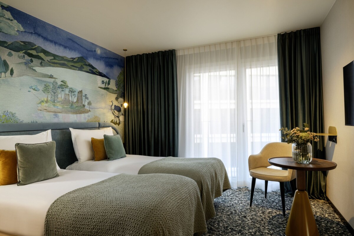 A hotel room with two beds, a chair, a small table, and a mural on the wall.