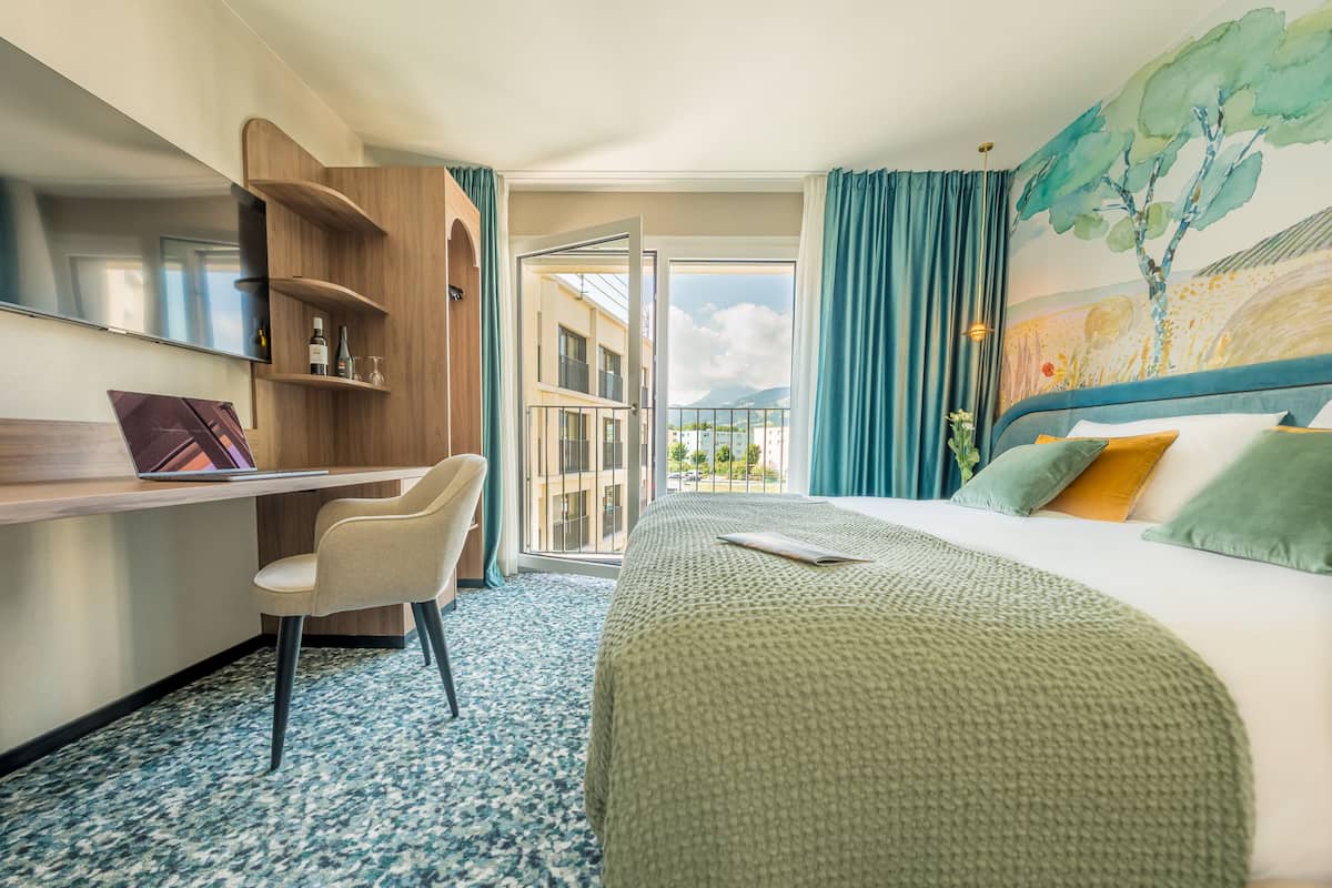 A hotel room with a bed, a desk, a chair, a TV, and a balcony with a view of buildings and mountains.