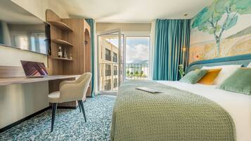 Ein Hotelzimmer mit Bett, Schreibtisch, Stuhl, Fernseher und einem Balkon mit Blick auf Gebäude und Berge.