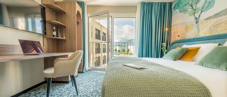 A hotel room with a bed, a desk, a chair, a TV, and a balcony with a view of buildings and mountains.
