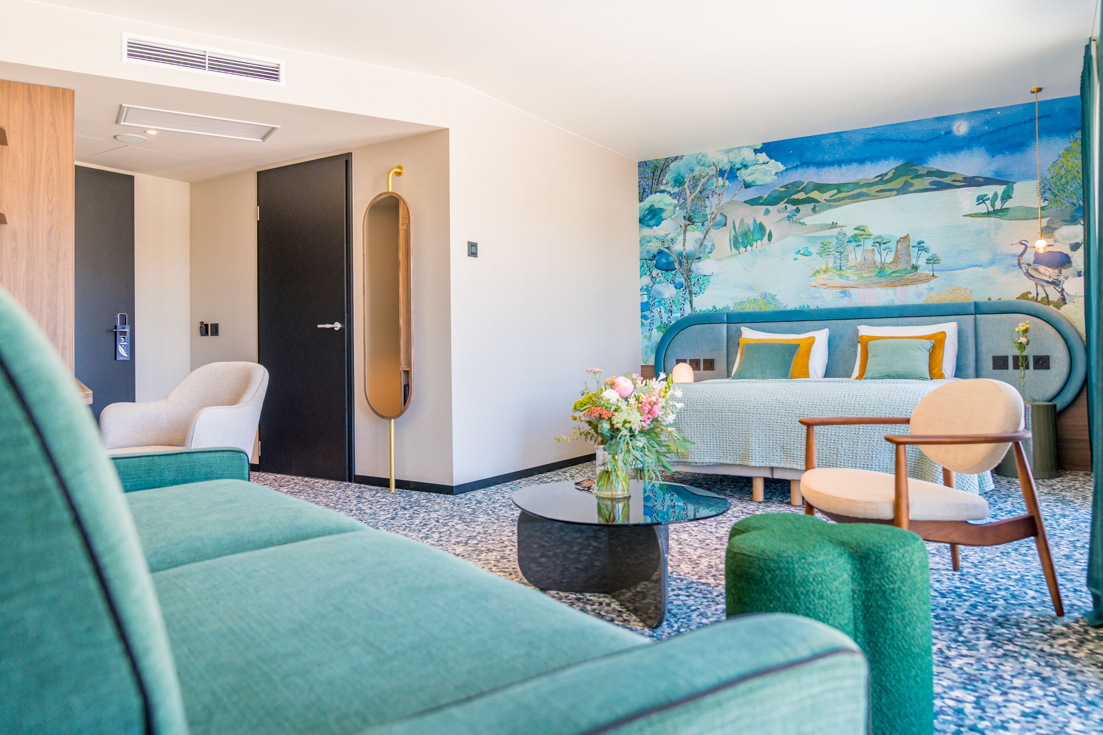Una habitación de hotel moderna con cama, dos sillones, una mesa de centro con flores y un mural en la pared.