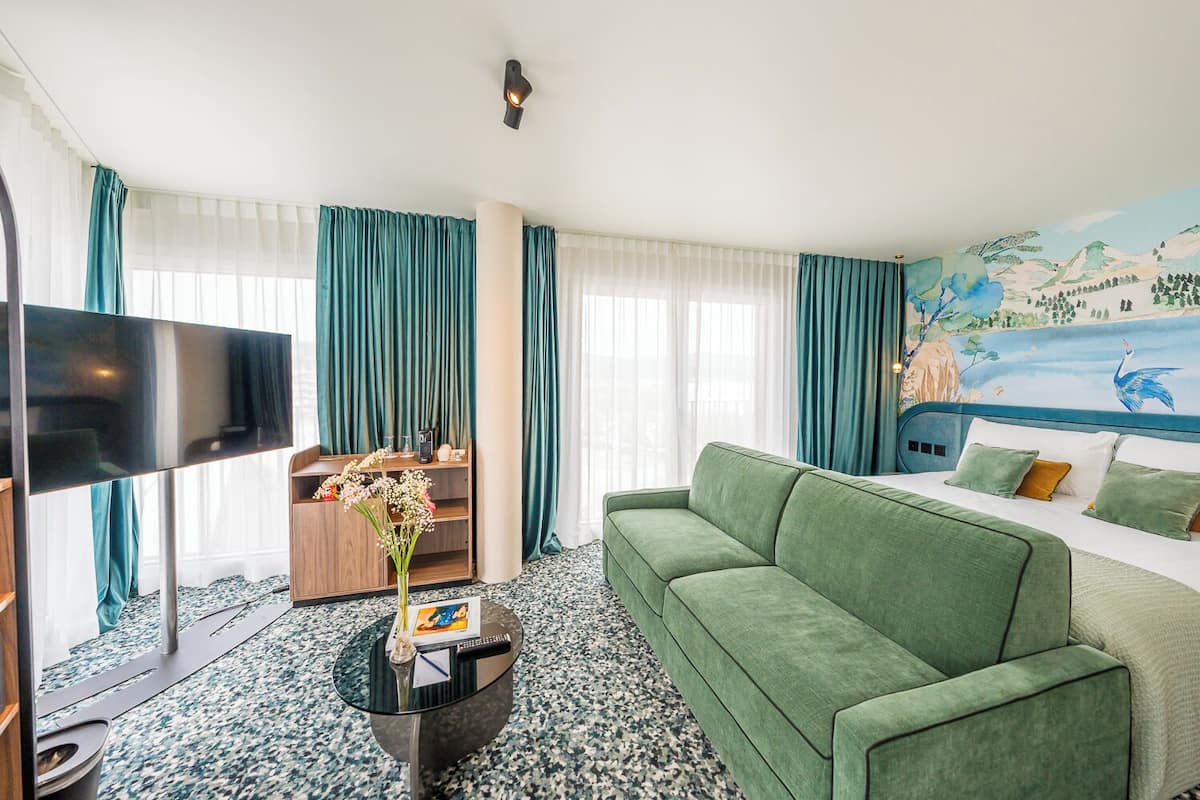 A hotel room with a green sofa, a TV, a small table with a vase of flowers, and a mural on the wall.