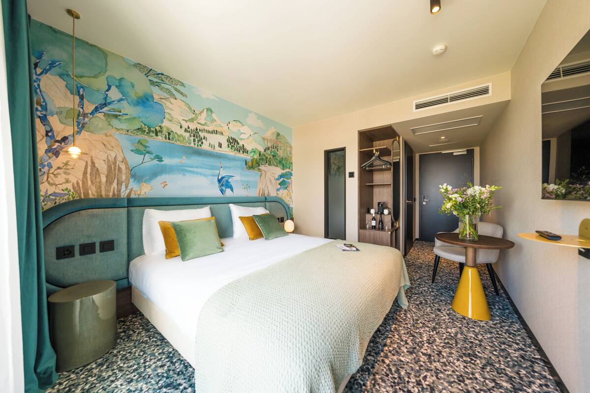 A hotel room with a bed, a wall mural, a round table, and a mirror.