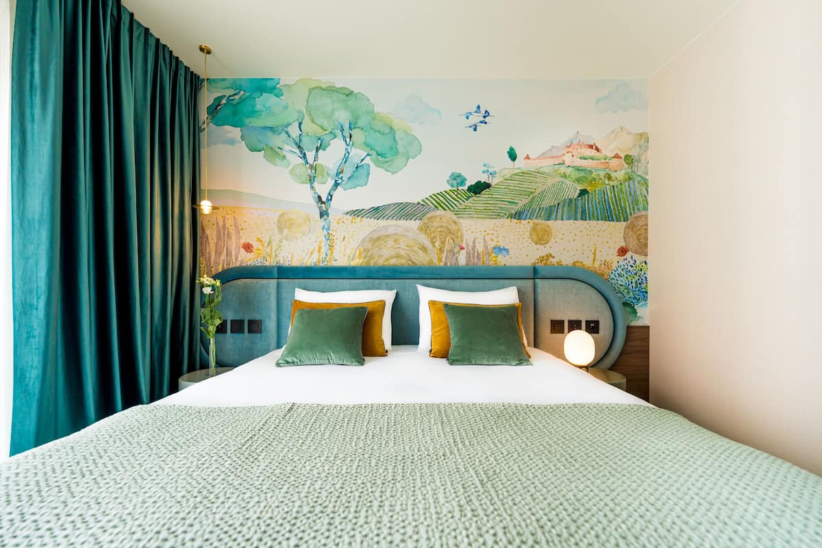 A neatly made bed with green and white pillows, a blue headboard, and a mural of a landscape on the wall behind.
