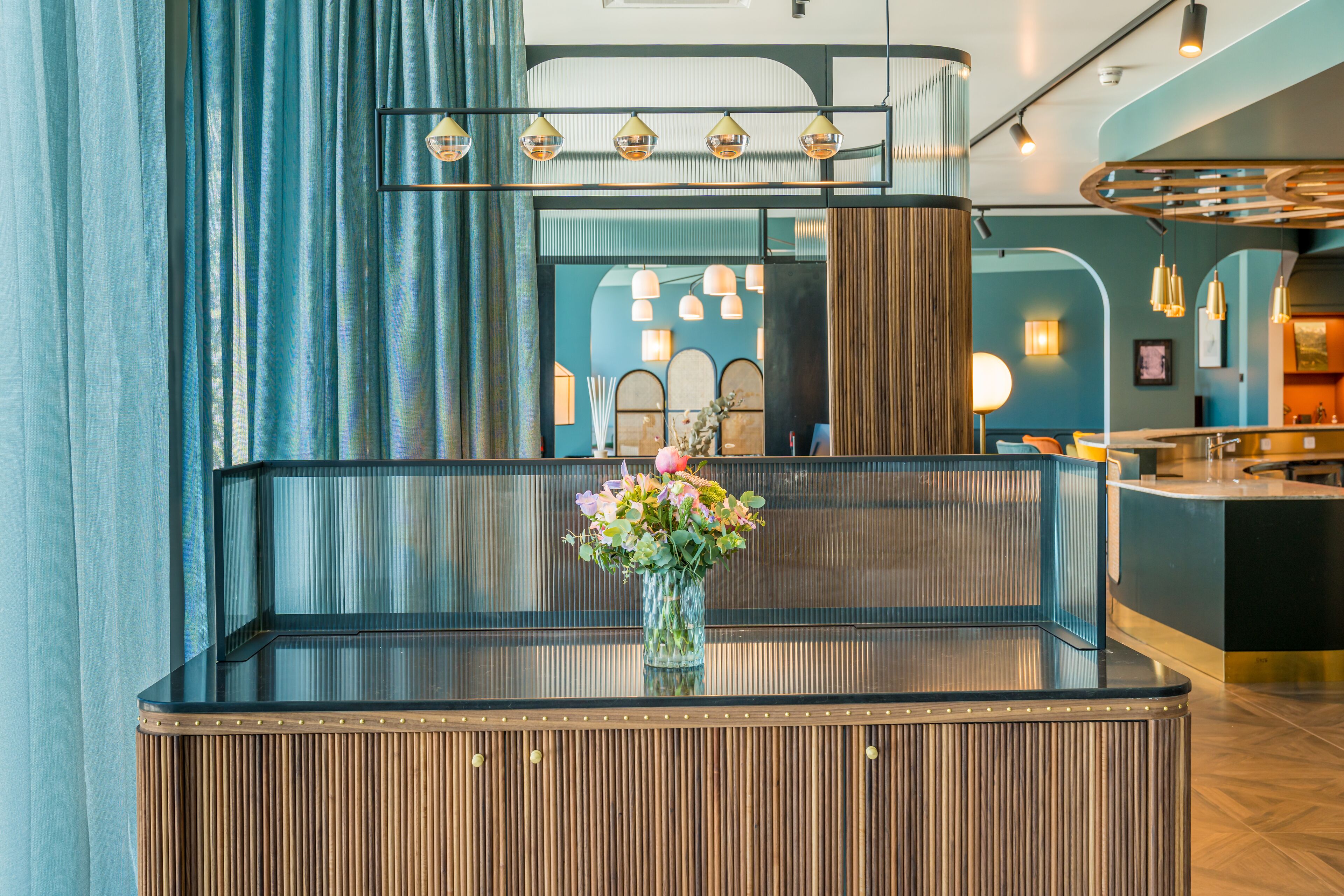 Een receptie met een bureau met glazen blad, een vaas met bloemen en een zithoek met een houten bank.