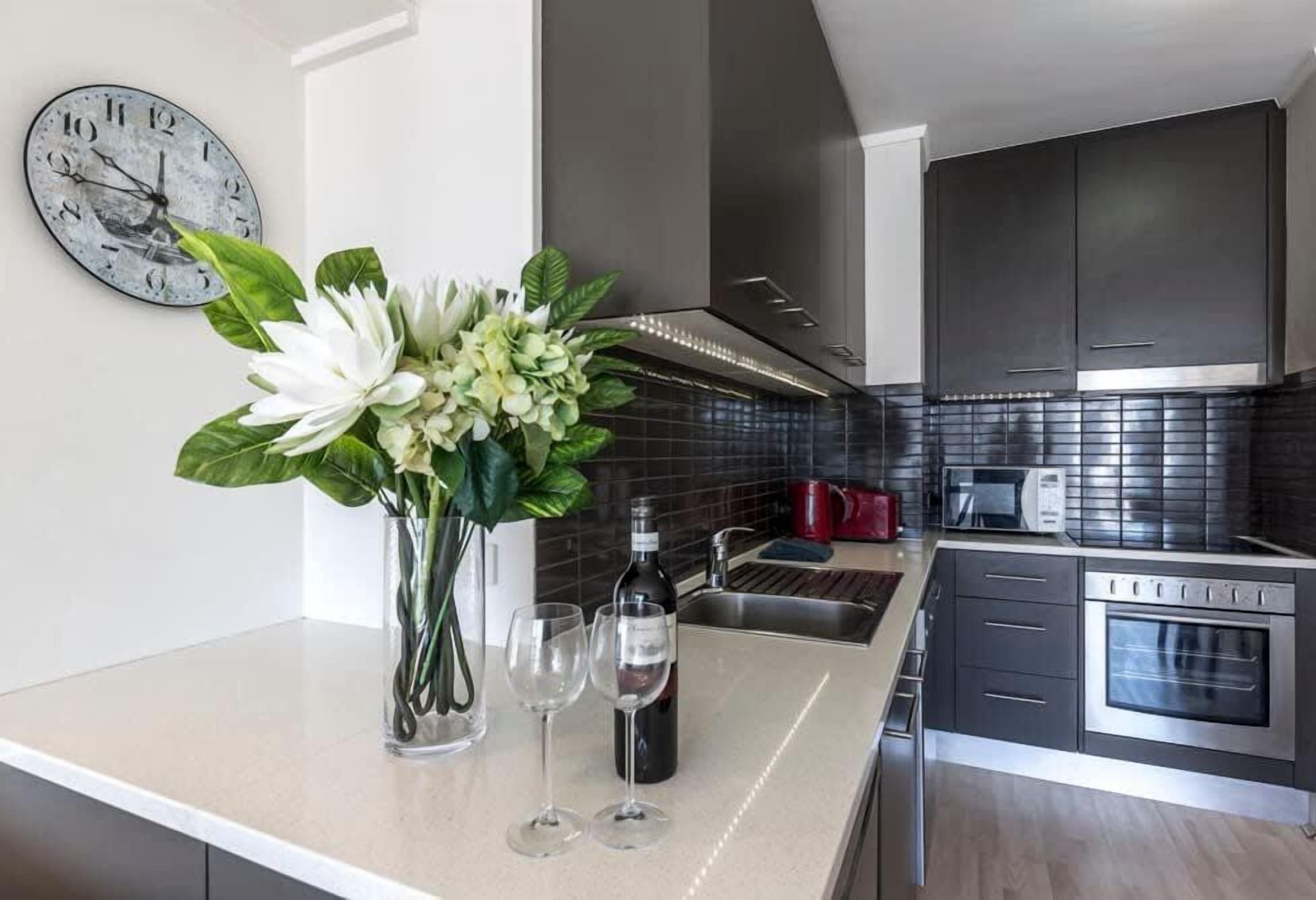 Une cuisine moderne avec un plan de travail blanc, des appareils en acier inoxydable, une horloge et un vase rempli de fleurs.