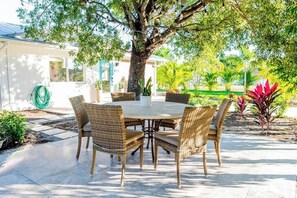 Een terras met een ronde tafel en rieten stoelen, omgeven door een boom en een tuin.