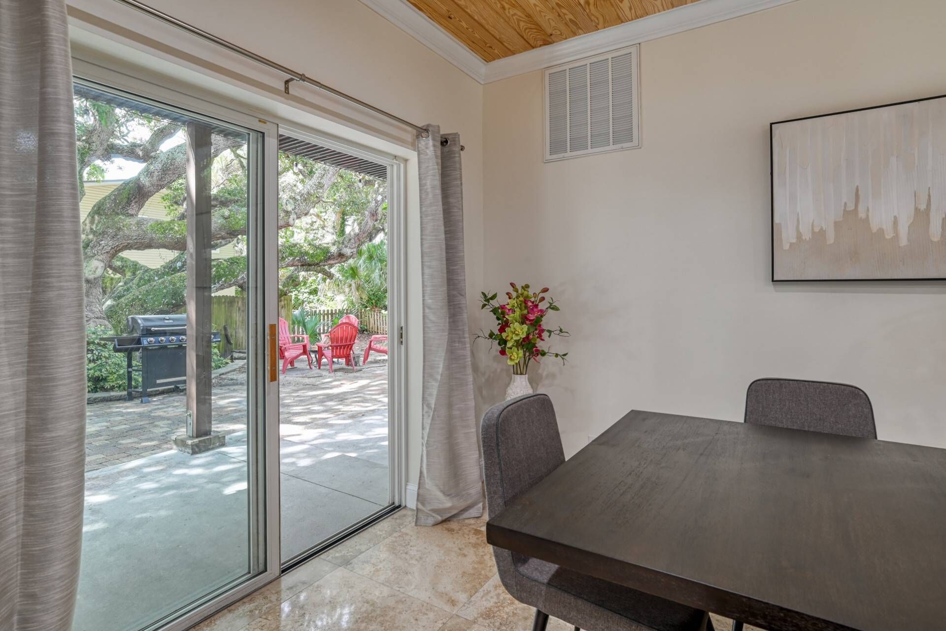 A room with a sliding glass door leading to a patio, a dining table with chairs, a vase with flowers, and a painting on the wall.