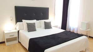 A neatly made bed with a black and white bedspread, two matching pillows, a bedside table with a lamp, and a wall-mounted light fixture. - ToledoRooms Palacios (Toledo)