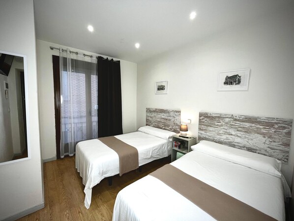 Una habitación de hotel con dos camas, una mesita de noche con lámpara, una ventana con cortinas y un espejo.