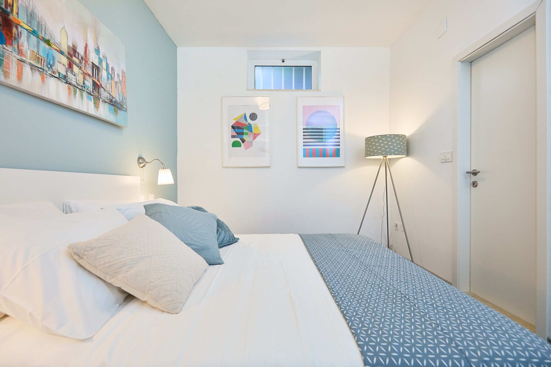A bedroom with a bed, pillows, a lamp, and two framed artworks on the wall.