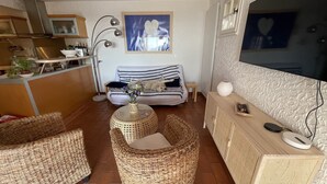 Un salon chaleureux avec des meubles en osier, un canapé à rayures, une petite table ronde ornée d’un vase et un téléviseur fixé au mur.