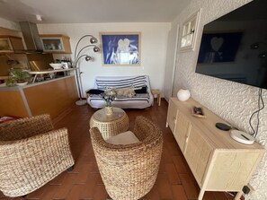 Een knusse woonkamer met rieten meubels, een gestreepte bank, een klein rond tafeltje met een vaas en een televisie aan de muur.