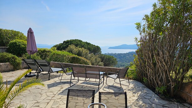 Un espace extérieur aménagé avec un sol en pierre, des tables et des chaises en bois, offrant une vue panoramique sur des collines et un plan d’eau.