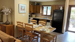 Uma área de refeições com uma mesa de madeira preparada para quatro pessoas, cercada por cadeiras de madeira. Uma cozinha é visível ao fundo, com armários de madeira e uma geladeira preta.