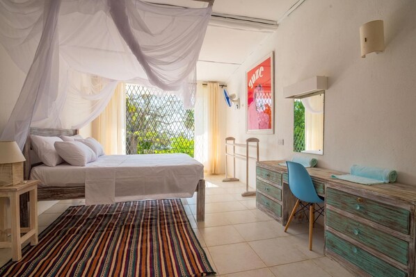A bedroom with a bed, a desk, a chair, and a window with a view of greenery.