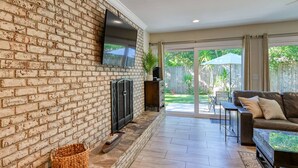 A living room with a brick wall, a flat-screen TV, a fireplace, a leather sofa, and a sliding glass door leading to a backyard. - La Valencia (St. Augustine)