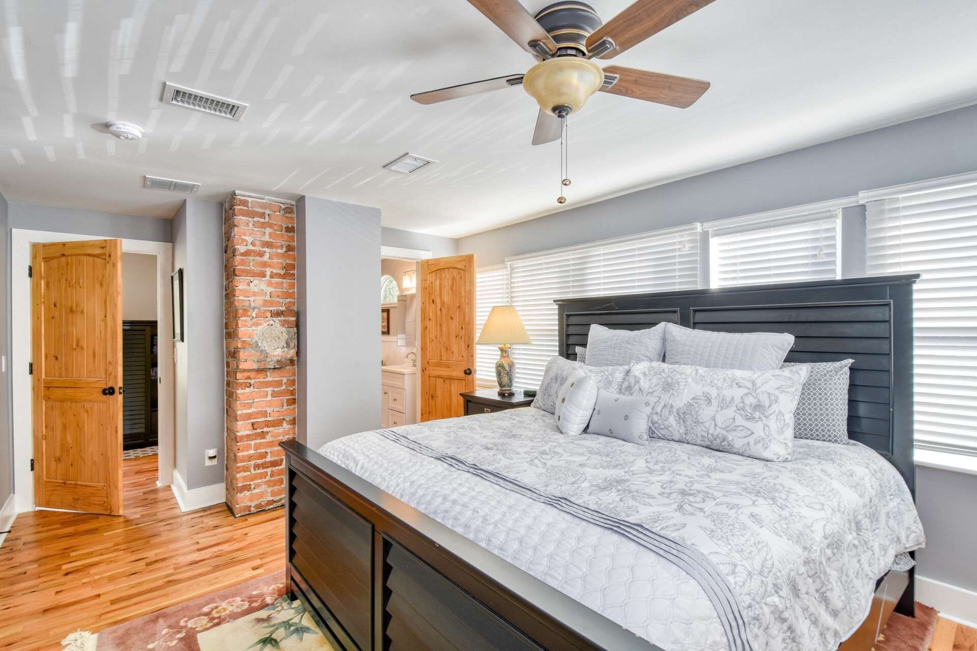 Une chambre avec un lit en bois, un ventilateur de plafond, une colonne en briques et une fenêtre avec des stores.
