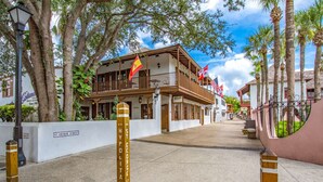Vista de la calle con un edificio que tiene balcones y banderas, rodeado de árboles y bollards.