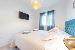 Une chambre à coucher avec un lit, un téléviseur fixé au mur, une fenêtre avec des rideaux et une lampe.