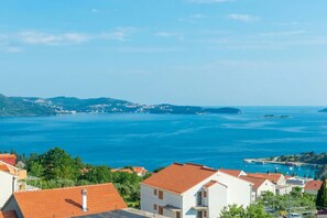 Une ville côtière aux maisons aux toits de tuiles rouges, dotée d’un port de plaisance et offrant une vue sur la mer et les collines lointaines.