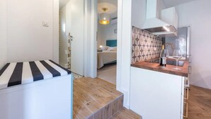 Una habitación de hotel moderna con una cama a rayas, suelo de madera, cocina con salpicadero estampado y un pasillo que lleva a otra habitación.