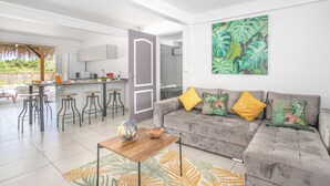 A modern living space with a grey sofa, a wooden coffee table, and a kitchen area with bar stools. - Villa Ajoupa (SAINTE ANNE)