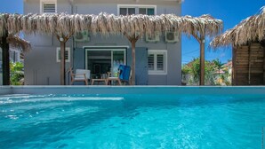 A swimming pool with a clear blue water, a wooden deck, and a building with a thatched roof and a door. - Villa Ajoupa (SAINTE ANNE)
