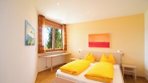 Une chambre à coucher comprenant un lit, deux tables de chevet, une fenêtre avec des rideaux, un radiateur et deux tableaux accrochés au mur.