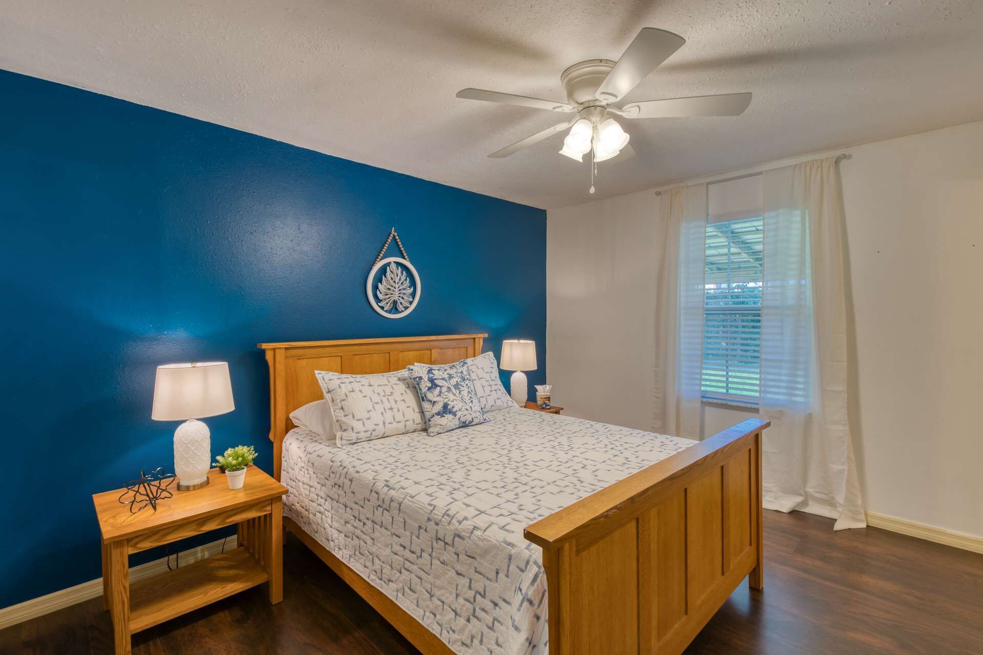 A bedroom with a wooden bed, two bedside tables with lamps, a ceiling fan, a window with blinds, and a decorative wall hanging.