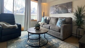 Ein Wohnzimmer mit Sofa, Sessel, Couchtisch und einem großen Fenster mit Vorhängen.