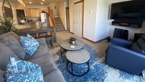 Ein modernes Wohnzimmer mit einem grauen Sofa, blauen Kissen mit Muster, einem Couchtisch und einem blauen Sessel. An der Wand ist ein Flachbildfernseher angebracht, und im Hintergrund ist eine Küche mit Holzschränken und einem Kühlschrank erkennbar.