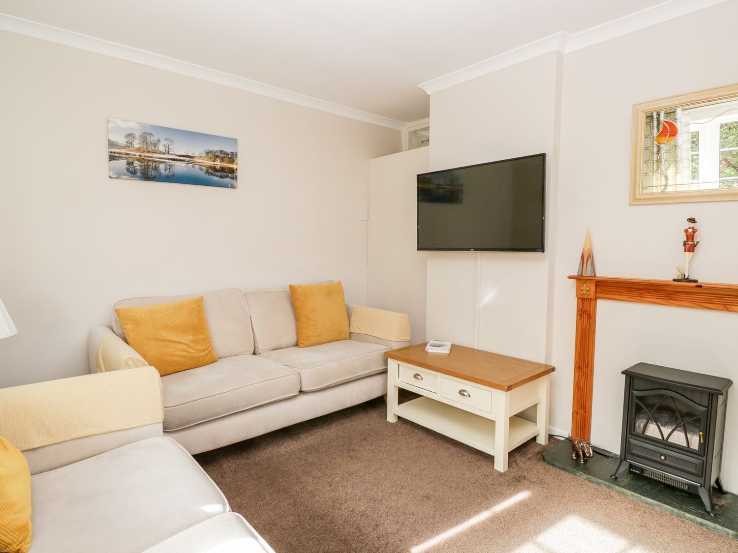 Een woonkamer met een beige bank, een klein houten tafeltje, een aan de muur bevestigde televisie, een open haard en een schilderij van een landschap.