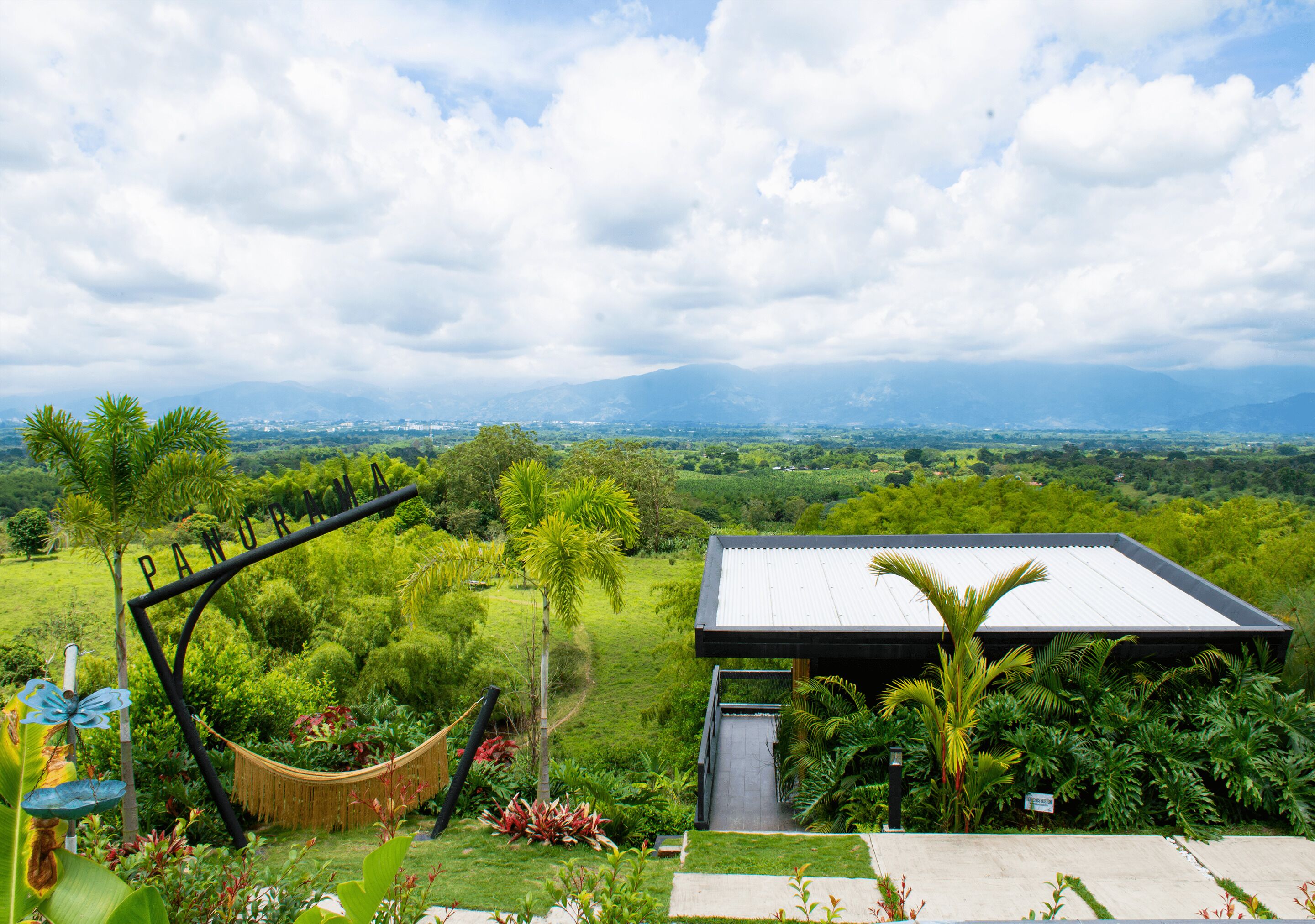 Un hamac dans un jardin luxuriant, avec vue sur des montagnes lointaines et un bâtiment doté d’un toit en métal.