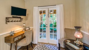 A living room with a television mounted on the wall, a wooden side table with a lamp, patterned chairs, a patterned rug, and a large glass door with curtains.