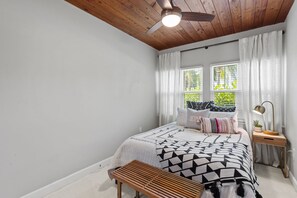 A bedroom with a bed, a bedside table with a lamp, a window with curtains, and a ceiling fan.