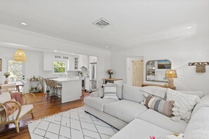 A modern, white-themed living room with a large sofa, a dining area with a table and chairs, and a kitchen with white cabinets and a central island.