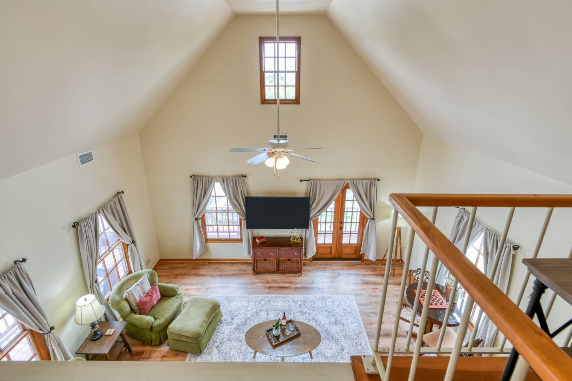 A spacious loft bedroom with a high ceiling, a ceiling fan, a large window, a bed, a desk, and a staircase.