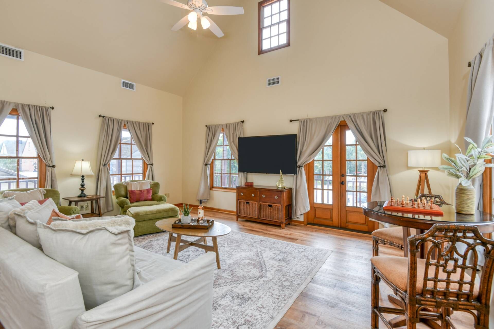 A spacious living room with a high ceiling, a flat-screen TV mounted on the wall, a green armchair, a white sofa, a wooden coffee table, and a dining table with chairs.