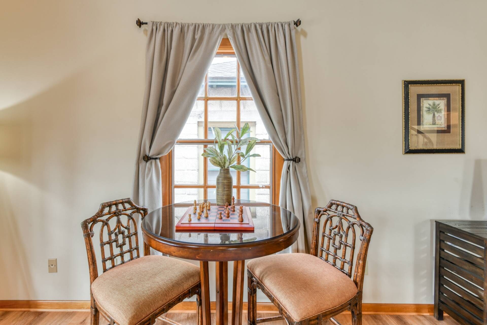 A dining area with a round table, two chairs, a vase with a plant, and a framed picture on the wall.