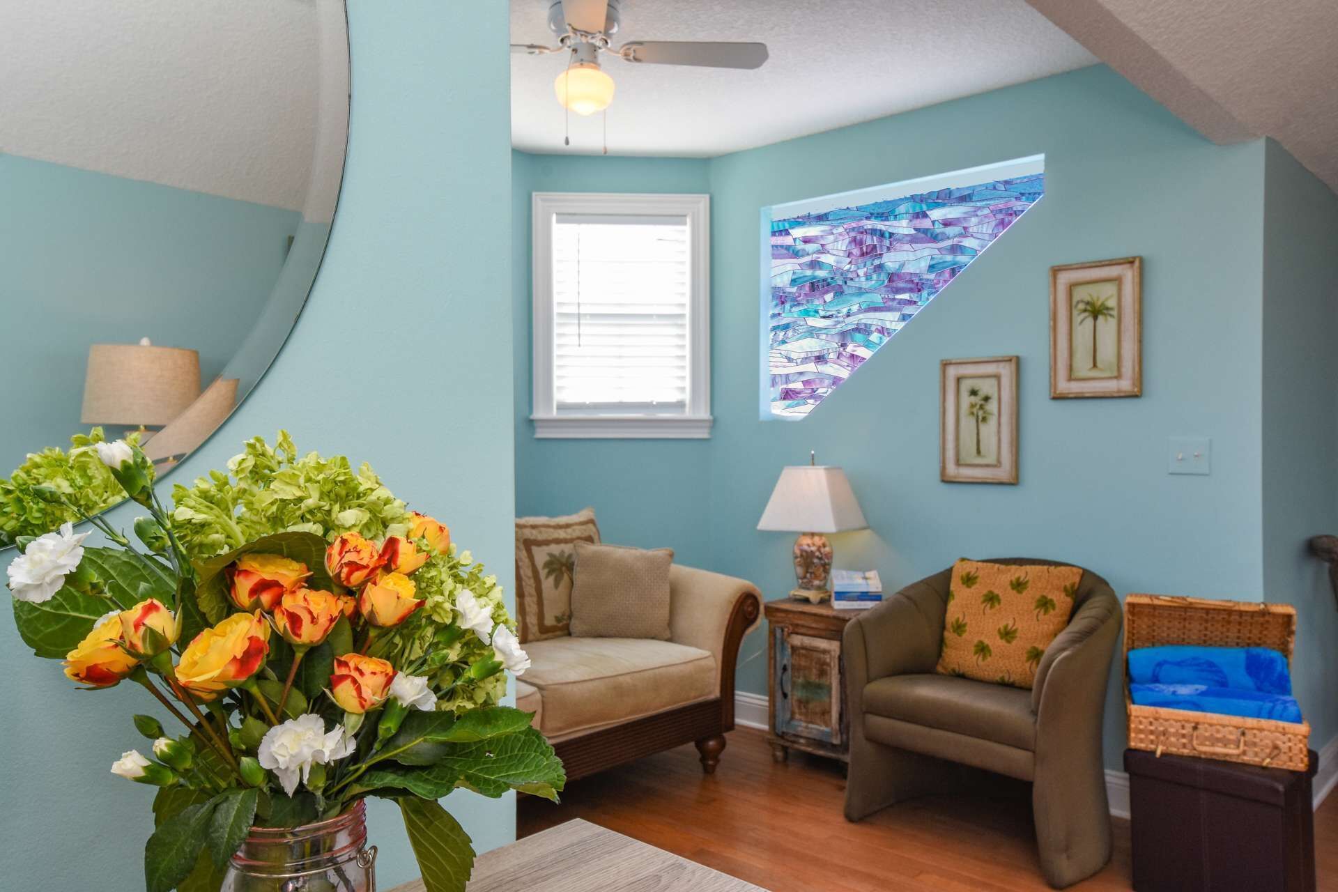 Un salon aux murs de couleur turquoise, doté d’un ventilateur de plafond, d’une fenêtre avec des stores, d’un vase de fleurs, de deux canapés, d’une table d’appoint avec une lampe et de tableaux encadrés accrochés au mur.