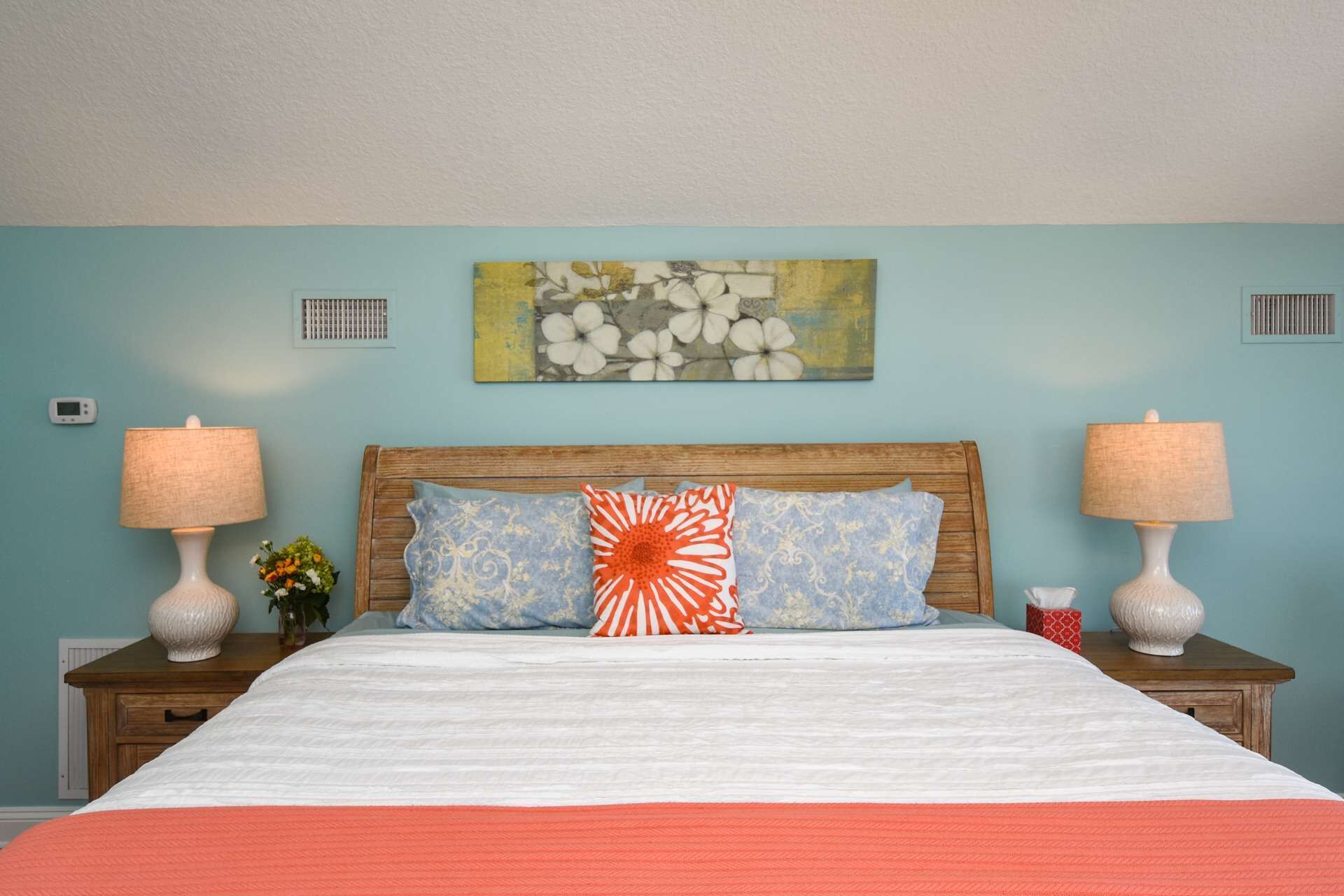 Habitación de hotel con cama, dos lámparas de noche, una mesita de noche con un florero y una obra de arte en la pared.