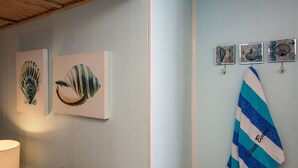 A room with two framed artworks on the wall, a surfboard leaning against the wall, and a lamp on a side table. - Seaside Studio (St. Augustine)